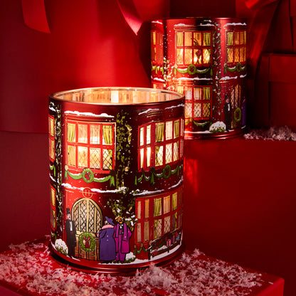 A cylindrical glass candle holder featuring a festive Christmas townhouse design with glowing windows, snow, green garlands, and holiday figures, set against a dark red background with a second matching tin behind it.